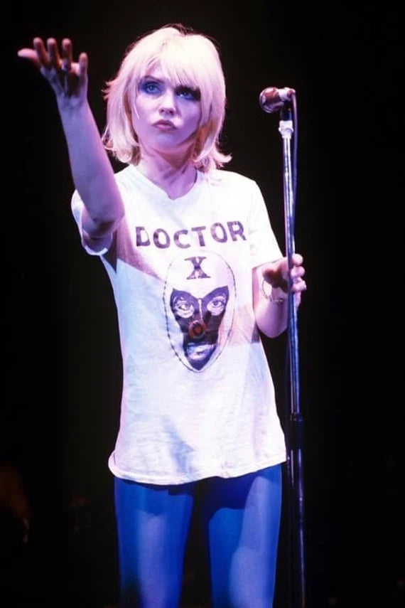 A woman with blonde hair is standing on stage, holding a microphone and gesturing with her hand. She is wearing a white t-shirt with a graphic design and the word "DOCTOR X" printed on it.