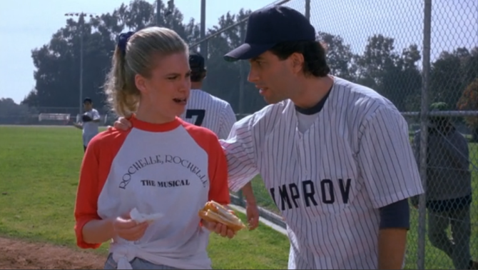 two people, a man and a woman, standing on a baseball field. The woman is wearing a red and white baseball jersey with the text "Rochelle, Rochelle THE MUSICAL" on it, and the man is wearing a baseball cap. They appear to be engaged in conversation or interaction, with the woman holding a hot dog.
