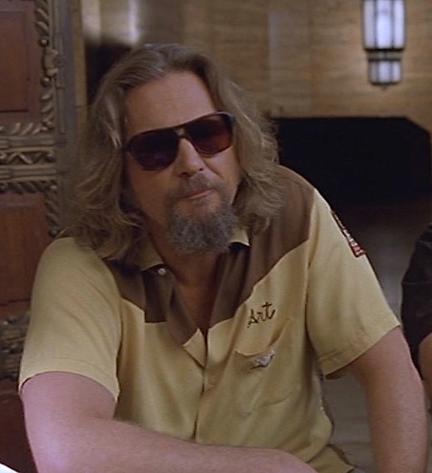 The Dude from the movie Big Lebowski sitting at a table wearing a brown and yellow bowling shirt with the name 'Art' on the front and sunglasses.
