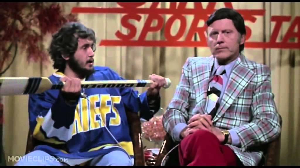 two men sitting in a chair, one wearing a blue and yellow sports jersey and the other wearing a plaid suit.