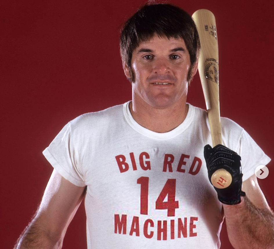 A man wearing a white t-shirt with the text "BIG RED 14 MACHINE" is holding a baseball bat and smiling.