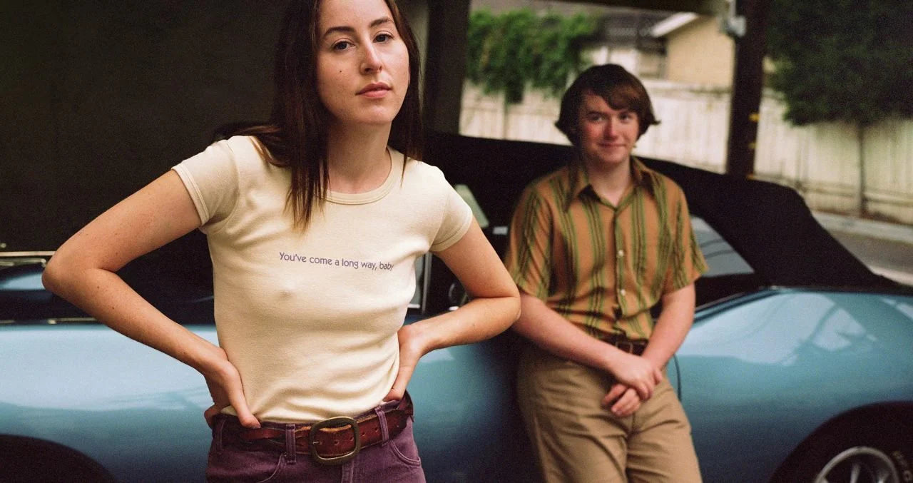A woman in a white t-shirt with the text "You've come a long way baby" stands next to a man in a striped shirt, both with their hands on their hips, in front of a blue car.