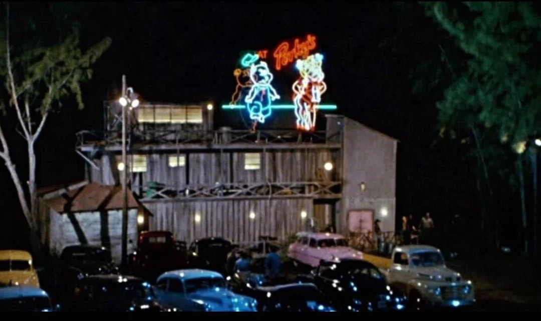 a neon sign for a bar or restaurant at night, with a large neon figure of a man and a woman on the sign. The scene is set in a parking lot with several cars parked around the area.
