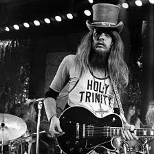 A black and white image of a man playing an electric guitar on stage, wearing a hat and a t-shirt with the text "HOLY TRINITY".