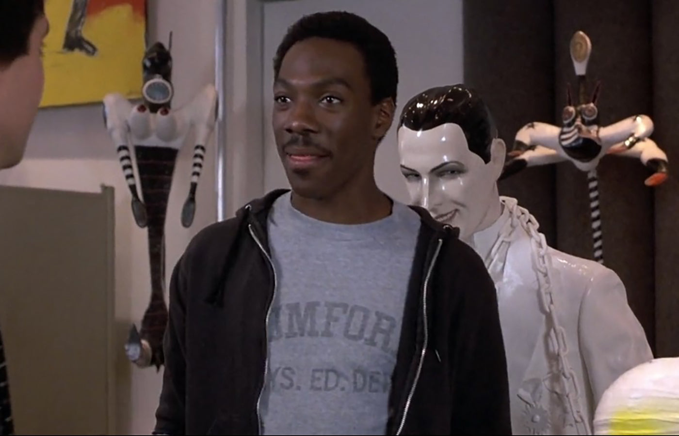 A man wearing a gray t-shirt with the text "COMFORT" is standing in an art gallery, looking at a sculpture of a man.