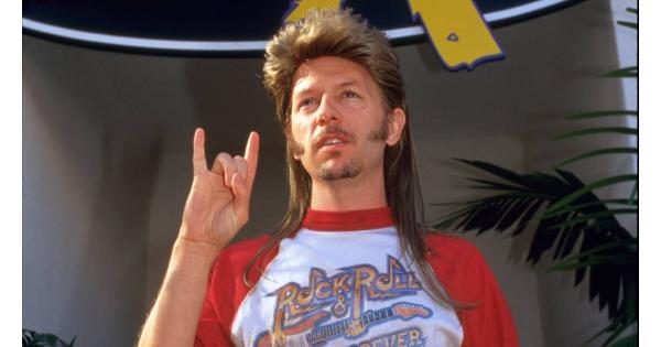 A man with long hair and a beard is making a hand gesture, wearing a red and white t-shirt with a graphic design.