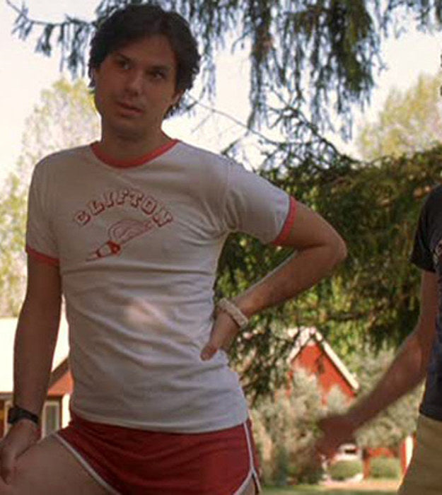 A man wearing a white t-shirt with a graphic and red shorts stands in front of a tree, with another person partially visible in the background.