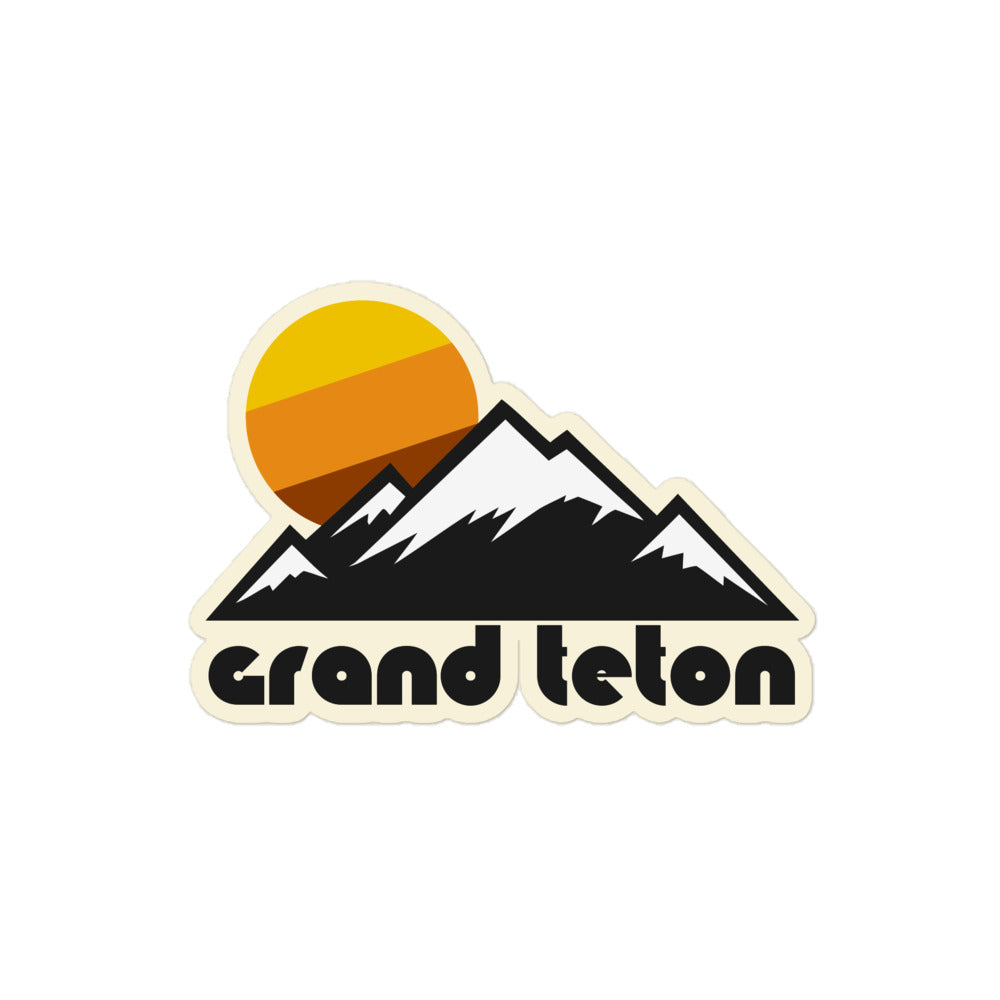A "Grand Teton" retro kiss cut sticker including the image of multiple pointed mountain peaks, snow covering them all, and a tricolored yellow/orange/brown sun in the distance.