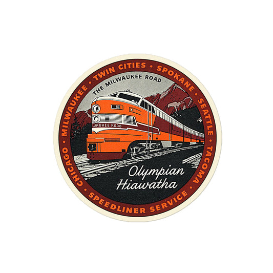 A circular retro Olympian Hiawatha kiss cut sticker that says "The Milwaukee Road" with the image of an orange and red train on gray tracks with the name 'Milwaukee Road', passing by red mountains and green forests and known as a "Speedliner Service". Kiss cut sticker surrounded by various cities including Tacoma, Seattle, Spokane, Twin Cities, MIlwaukee, and Chicago which the train would visit from 1847-1986.