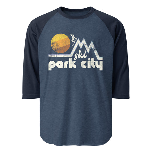 A 3/4 sleeve heather blue and navy colored raglan shirt that says "Ski Park City", referencing the famous skiing town in Utah with Park City Mountain Resort and Deer Valley Resort. Includes image of a skier headed down one of many mountain slopes, passing by a beautiful setting sun.