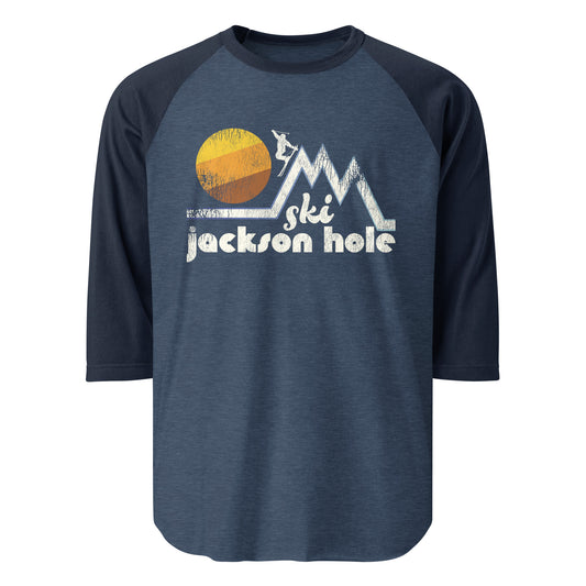 A vintage 3/4 sleeve navy and heather blue raglan shirt saying "Ski Jackson Hole" under multiple triangular mountains and a person skiing down as a yellow, orange, and brown colored sun set.