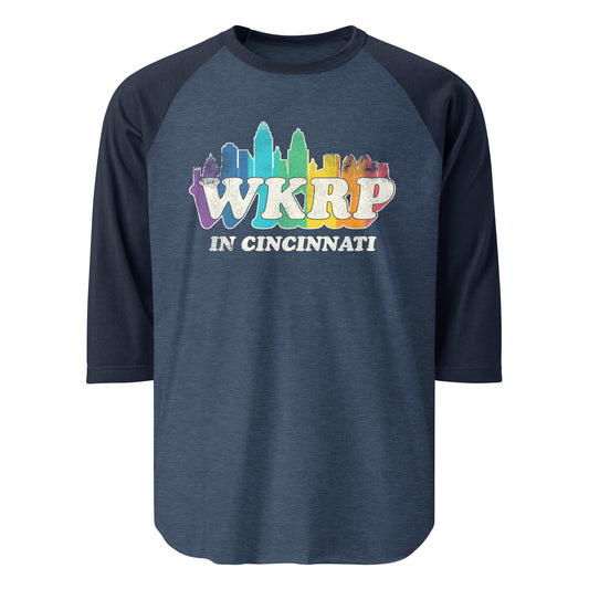 An 80s raglan style three quarter sleeve heather blue and navy WKRP in Cincinnati navy shirt. Has the Cincinnati, Ohio skyline in rainbow colors. References the comedy TV series from 1978-1982.