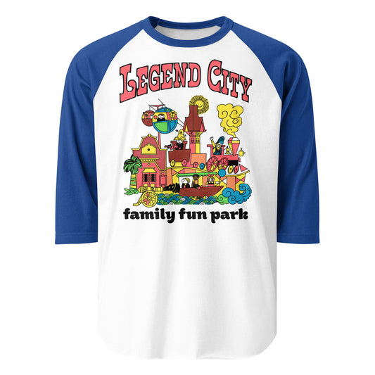 A tourist blue and white three quarter sleeve raglan shirt that says "Legend City family fun park" with images of children riding trains, skyway gondolas, boats and more in the Arizona amusement park. Images of a smiling sun, a cannon with the word "boom", a palm tree, and family friendly cute buildings.