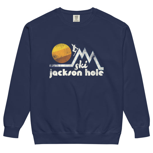 A navy colored "Ski Jackson Hole" retro crew neck sweatshirt for tourists. Has image of a skier jumping down a steep mountain slope as a yellow, orange, and brown sun sets. References the Wyoming city popular for skiers, snowboarders, and hikers in the Teton Mountains.