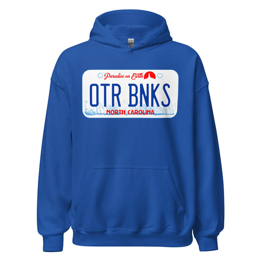 A royal blue Outer Banks retro 70s style hooded sweatshirt with the image of a car license plate with that says "OTR BNKS", "North Carolina", and "Paradise on Earth" in front of light blue American beachgrass swaying in the wind a tiny lighthouse circular red icon. References the Netflix 2020 5 season TV Series that takes place in OBX.