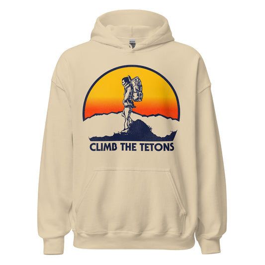 A sand colored 80s style tourist hoodie sweatshirt with a hiker at the top of a mountain, hands in pockets, as a huge sun sets. Words say "Climb the Tetons", referencing the famous national park The Grand Tetons, located near Jackson, Wyoming and Yellowstone National Park.