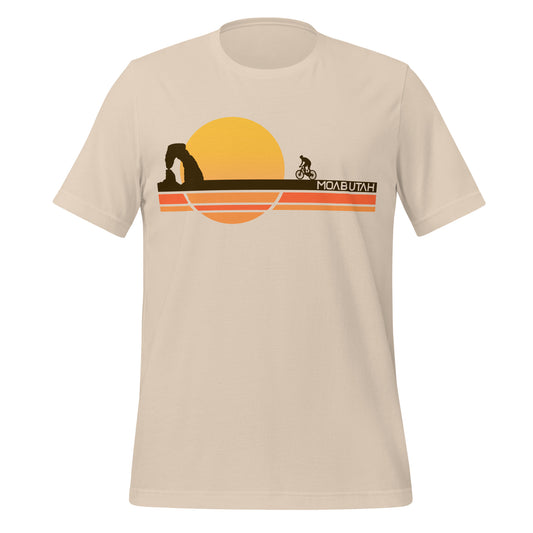 An 80s style soft cream colored Moab, Utah t-shirt for tourists with stripes of orange, yellow, and brown land over which a person rides a bike, and a sun setting. The iconic landmark Delicate Arch stands in shadow in the distance, the most famous natural stone arch in the world, a part of Arches National Park.