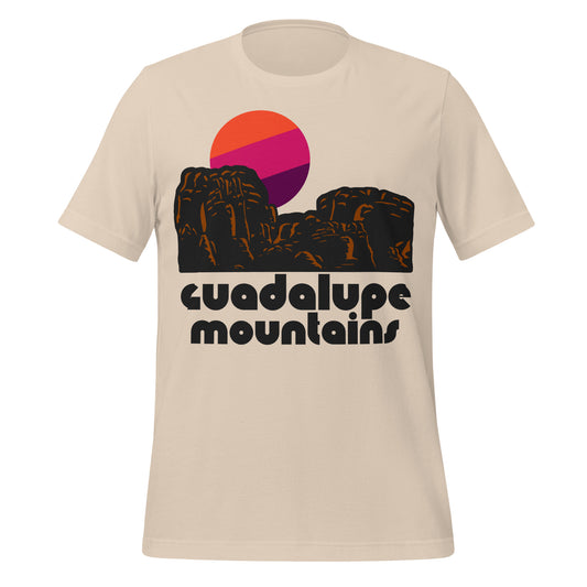 A 70s style soft cream short sleeve t-shirt for tourists with the words "Guadalupe Mountains" in front of red rock mountains and an orange, pink, and magenta sun.
