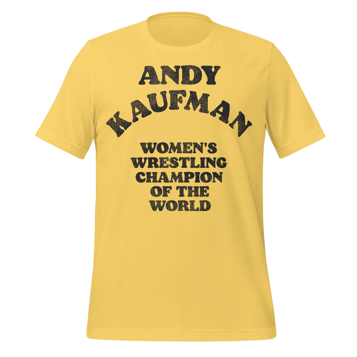 A yellow retro 80s style t-shirt that says "Andy Kaufman", "Women's Wrestling Champion of the World". From the 2025 biopic movie Queen of the Ring about Mildred Burke directed by Ash Avildsen.