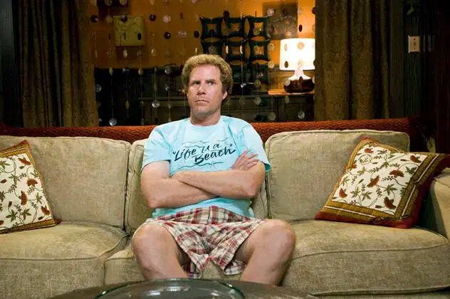 Will ferrell as Brennan in the movie Step Brothers sitting on a coach with his arms crossed wearing 'Life is a Beach' blue t shirt.