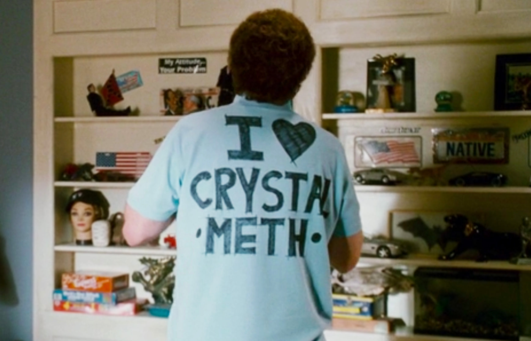 A person wearing a light blue t-shirt with the text "I ♥ CRYSTAL METH" is standing in front of a bookshelf filled with various items.
