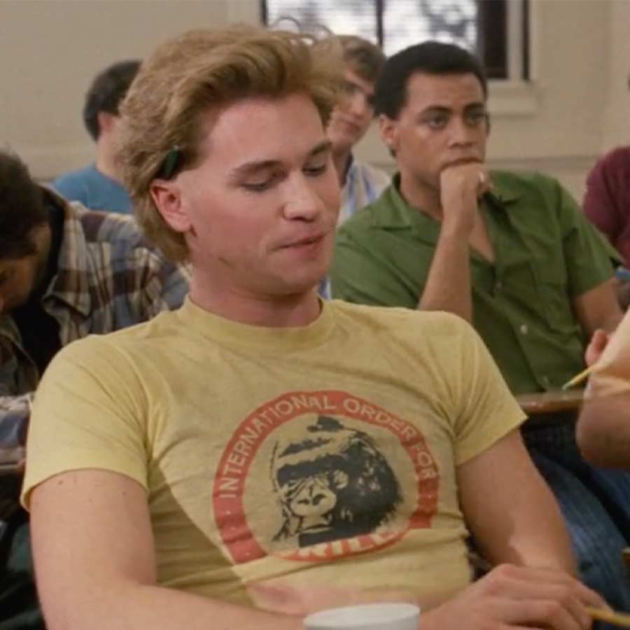 A man wearing a yellow t-shirt with a graphic of a gorilla is sitting in a classroom, looking down.