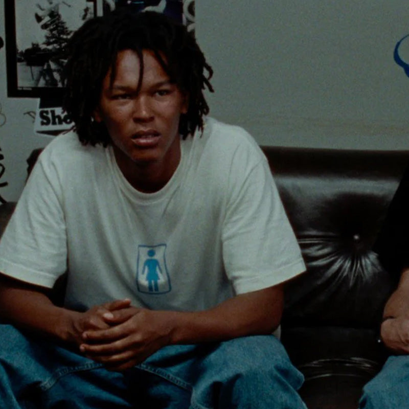 A young man with dreadlocks is sitting on a leather couch, wearing a white t-shirt with a blue graphic.