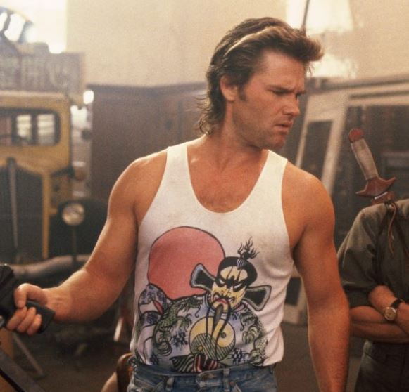 A man wearing a white tank top with a graphic design is standing in a room with various objects, including a yellow truck and a motorcycle.