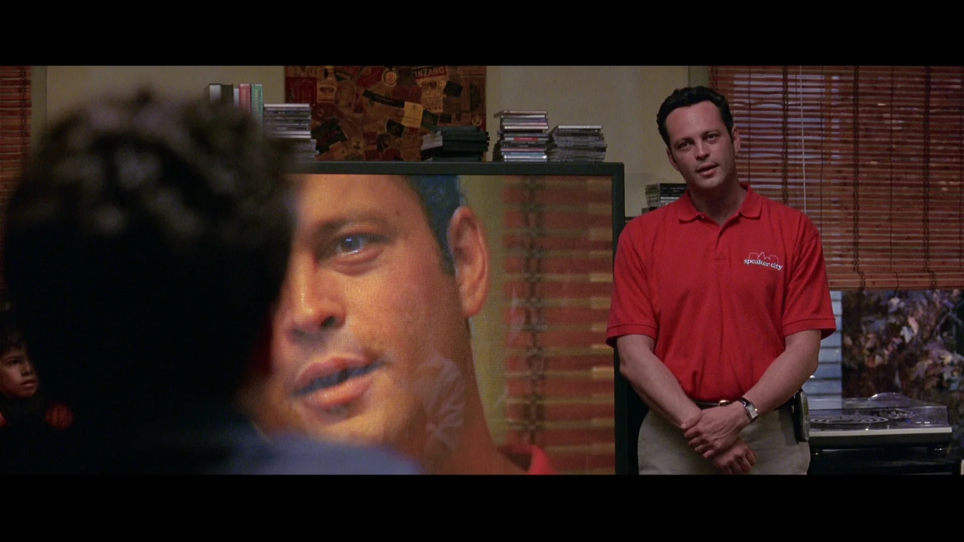a man in a red shirt standing in a room, with a large screen displaying a close-up of another man's face.