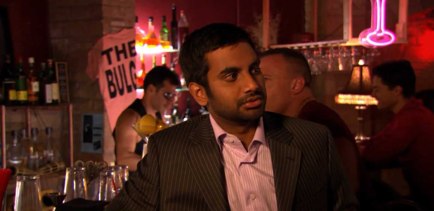 A man in a suit sits at a bar, surrounded by bottles and glasses, with a neon sign in the background.