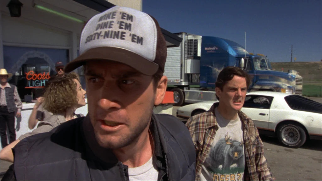 two men standing in front of a truck, with one of them wearing a baseball cap with text on it.