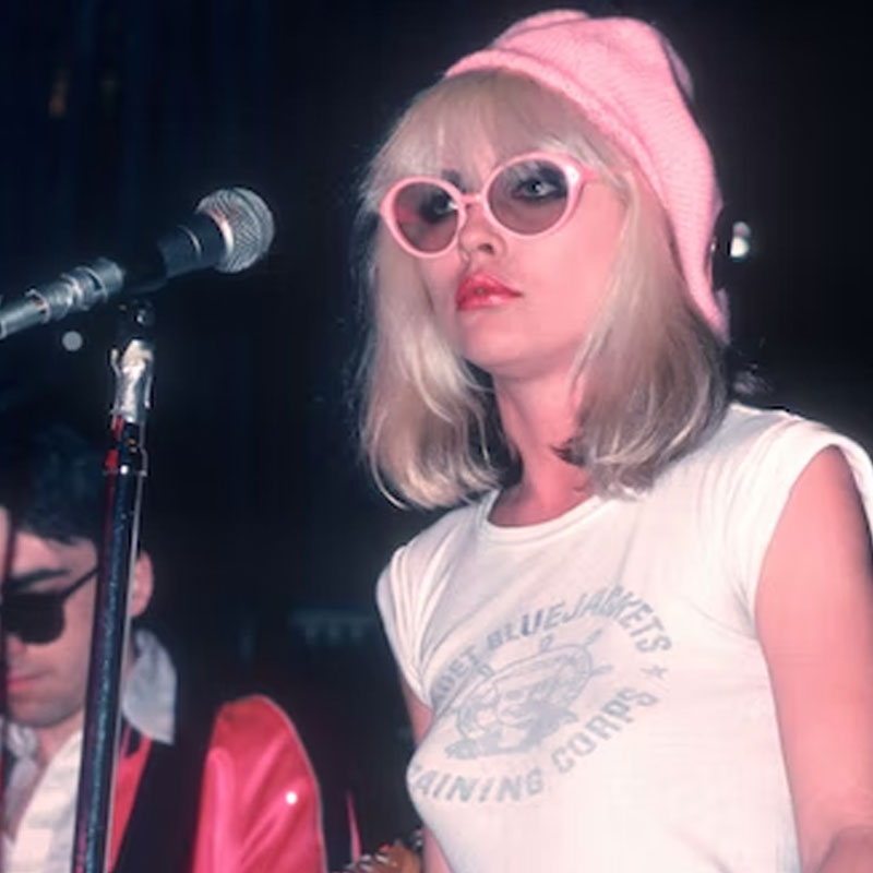 A woman wearing a pink beanie and sunglasses is performing on stage, holding a microphone and wearing a white t-shirt with text.