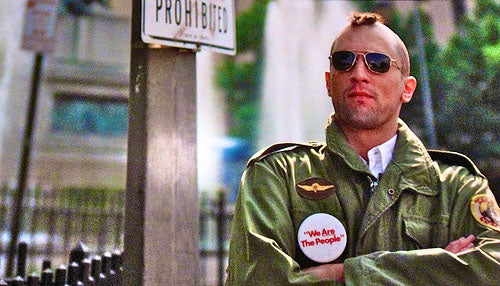 A man in a green military jacket stands with his arms crossed, wearing sunglasses and a "We're Art The People" button.