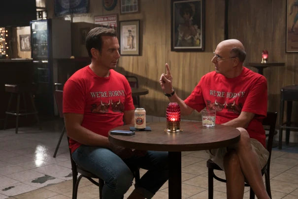 Two men wearing red t-shirts with the text "We're Born" are seated at a table in a dimly lit bar, engaged in conversation.