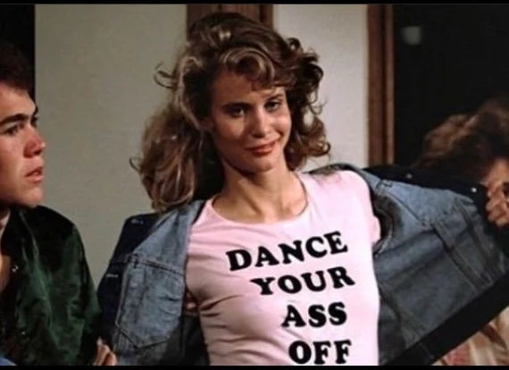 A woman with curly hair wearing a pink t-shirt with the text "DANCE YOUR ASS OFF" is smiling and looking at the camera.