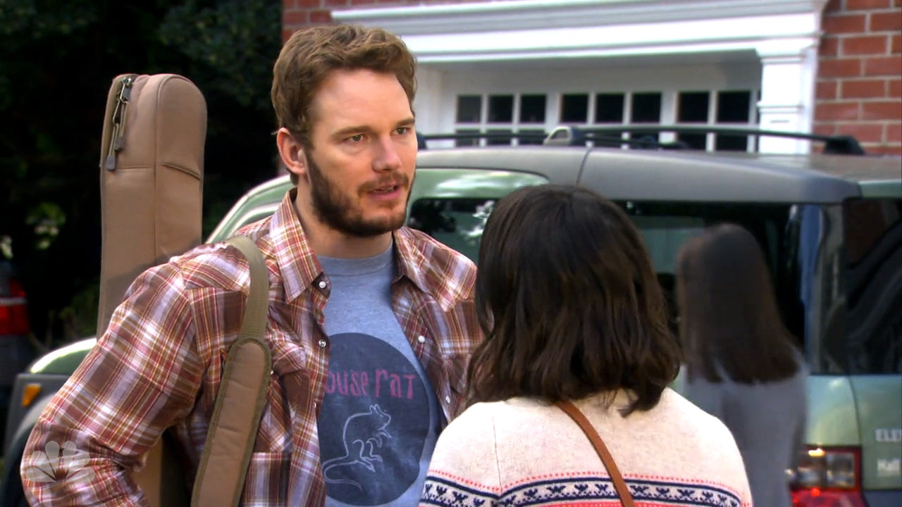 A man in a plaid shirt and a woman in a sweater are standing in front of a parked car, engaged in conversation.