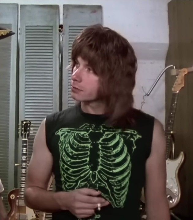 A man with long hair wearing a black sleeveless shirt with a green skeleton design stands in a room with various musical instruments.