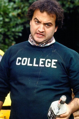 A man wearing a blue sweater with the word "COLLEGE" on it, holding a bottle of alcohol and making a facial expression.