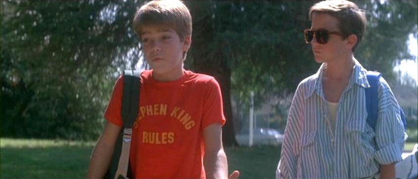 two young boys walking together in a park, with one wearing a red t-shirt and the other wearing sunglasses.