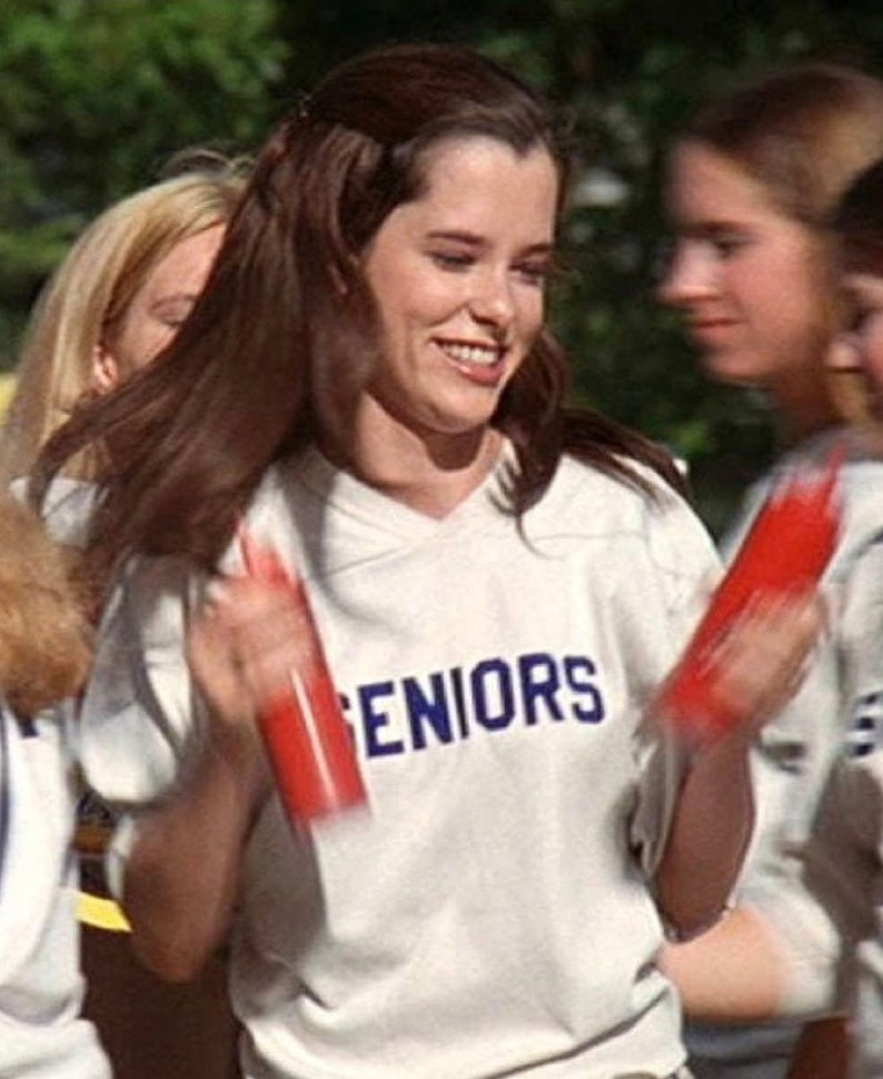 A young woman with long brown hair is smiling and holding a red object, wearing a white shirt with the word "GENIORS" written on it.