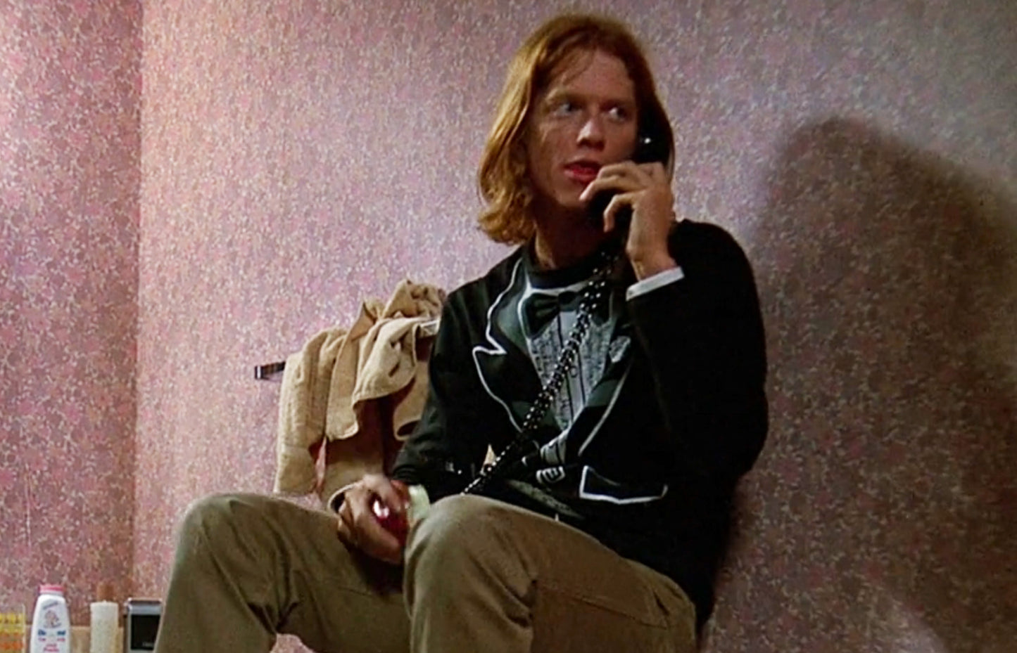 A man with long hair is sitting in a room, talking on a phone, and holding a bottle in his hand.