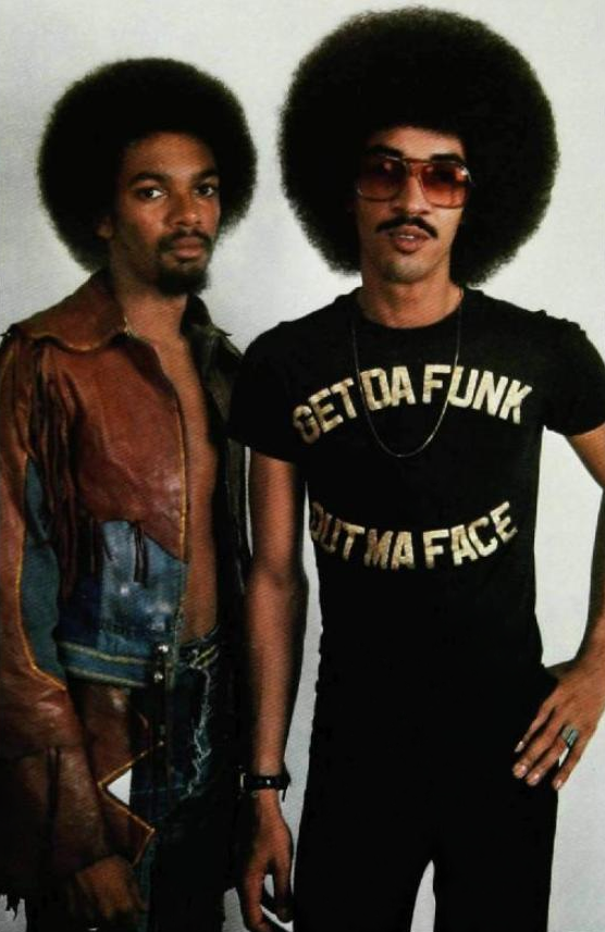 Two men with large afro hairstyles and sunglasses pose together, one wearing a leather jacket and the other wearing a black t-shirt with the text "GET DA FUNK OUT MA FACE".