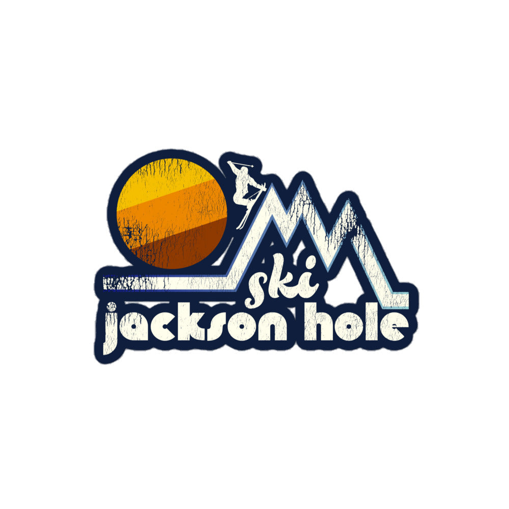 A vintage retro "Ski Jackson Hole" kiss cut sticker that includes the image of a skier jumping down a steep mountain peak in front of a yellow, orange, and brown setting sun. 