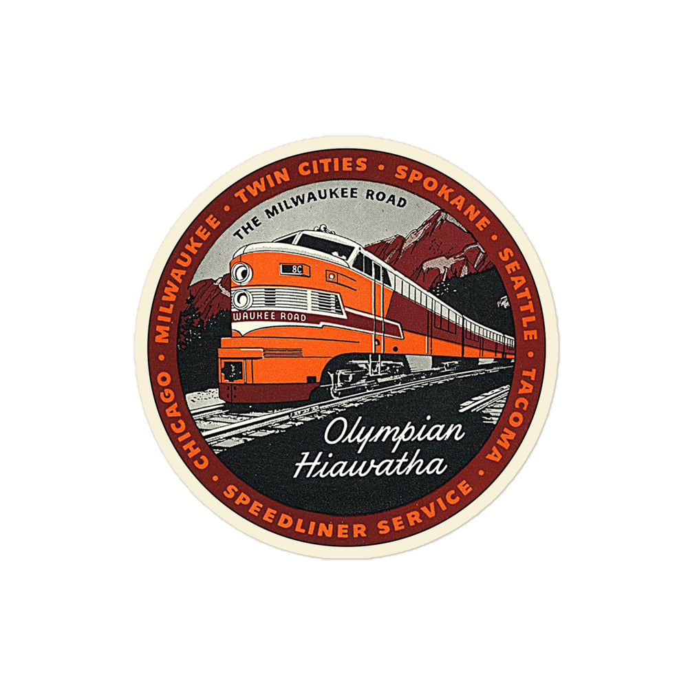 A circular retro Olympian Hiawatha kiss cut sticker that says "The Milwaukee Road" with the image of an orange and red train on gray tracks with the name 'Milwaukee Road', passing by red mountains and green forests and known as a "Speedliner Service". Kiss cut sticker surrounded by various cities including Tacoma, Seattle, Spokane, Twin Cities, MIlwaukee, and Chicago which the train would visit from 1847-1986. 