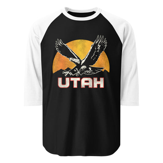 A 3/4 sleeve white and black raglan style Utah shirt with the image of a golden eagle, considered Utah's state bird of prey, in front of a sun setting over a mountain range. 