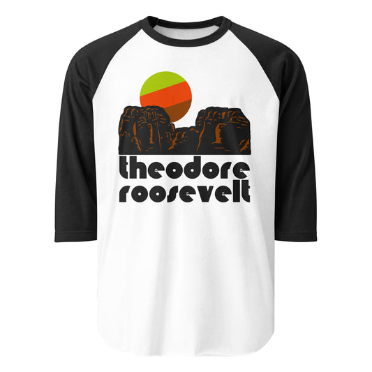 A black and white 3/4 sleeve retro raglan style shirt that says "Theodore Roosevelt' in reference to the national park named after him under an image of a rocky Badlands landscape and a green/red/brown sun. 