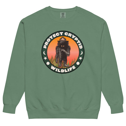 A green crew cut retro Protect Cryptid Wildlife sweatshirt. Has image of unknown beast with black fur and yellow eyes inside a forest with a pink and orange sunset. Has symbols of a sun, star, and the moon. 