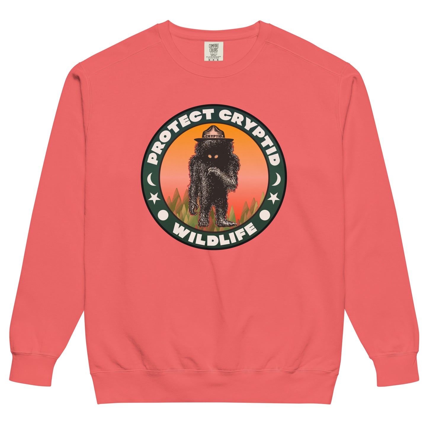 A watermelon pink colored retro crew neck sweatshirt with the words Protect Cryptid Wildlife and the image of an undiscovered black and hairy creature with yellow eyes, inside a forest against a pink and orange backdrop. Has a hat labeled "cryptid."