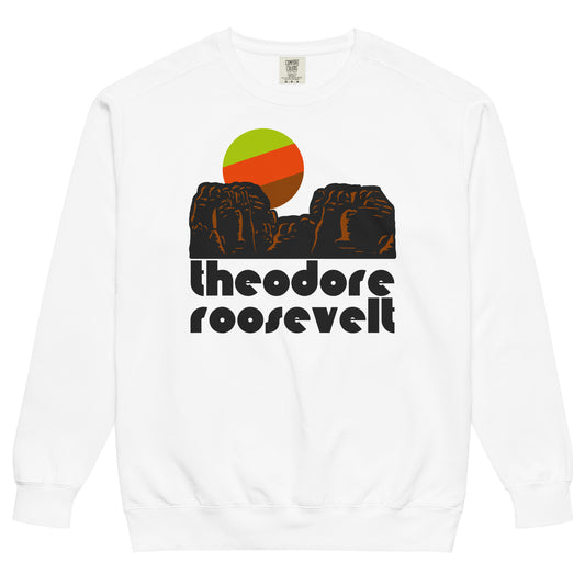 A retro, 80s style souvenir "Theodore Roosevelt" National Park white crew neck sweatshirt. Includes the image of stylized reddish brown rocks, resembling the Badlands landscape, and a setting tricolored sun. Located in North Dakota, the park is at the intersection of the Badlands and the Great Plains and includes the Little Missouri River, horse and hiking trails, and wildlife like bison. 