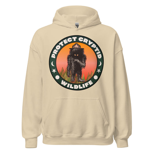 A vintage sand colored Protect Cryptid Wildlife hoodie sweatshirt with the image of an a black, hairy beast with yellow eyes, on top of a green forest against a pink and orange sunrise with a hat labeled Cryptid. 